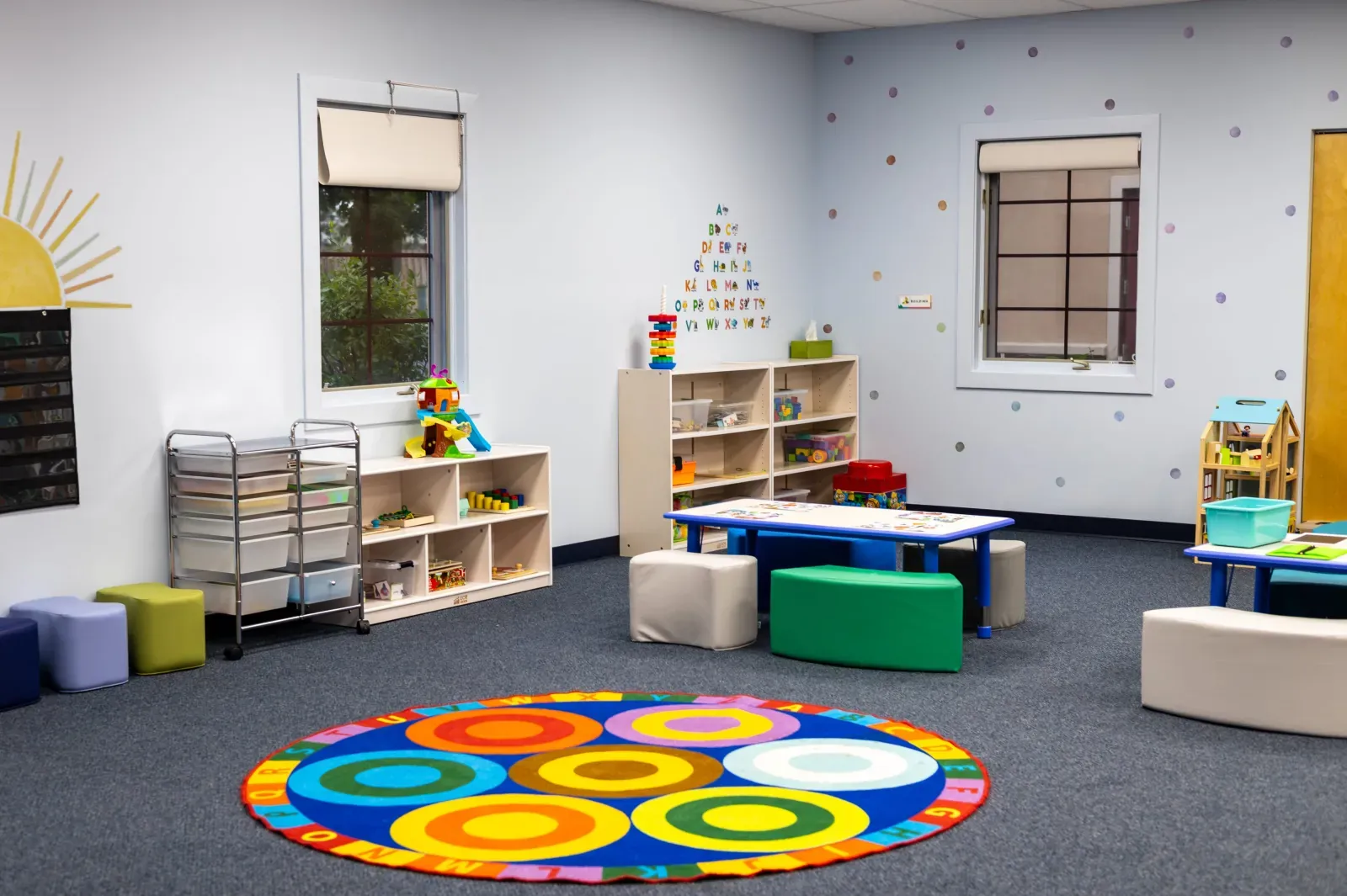 Therapy classroom with a colorful circle rug, small tables and toy shelves